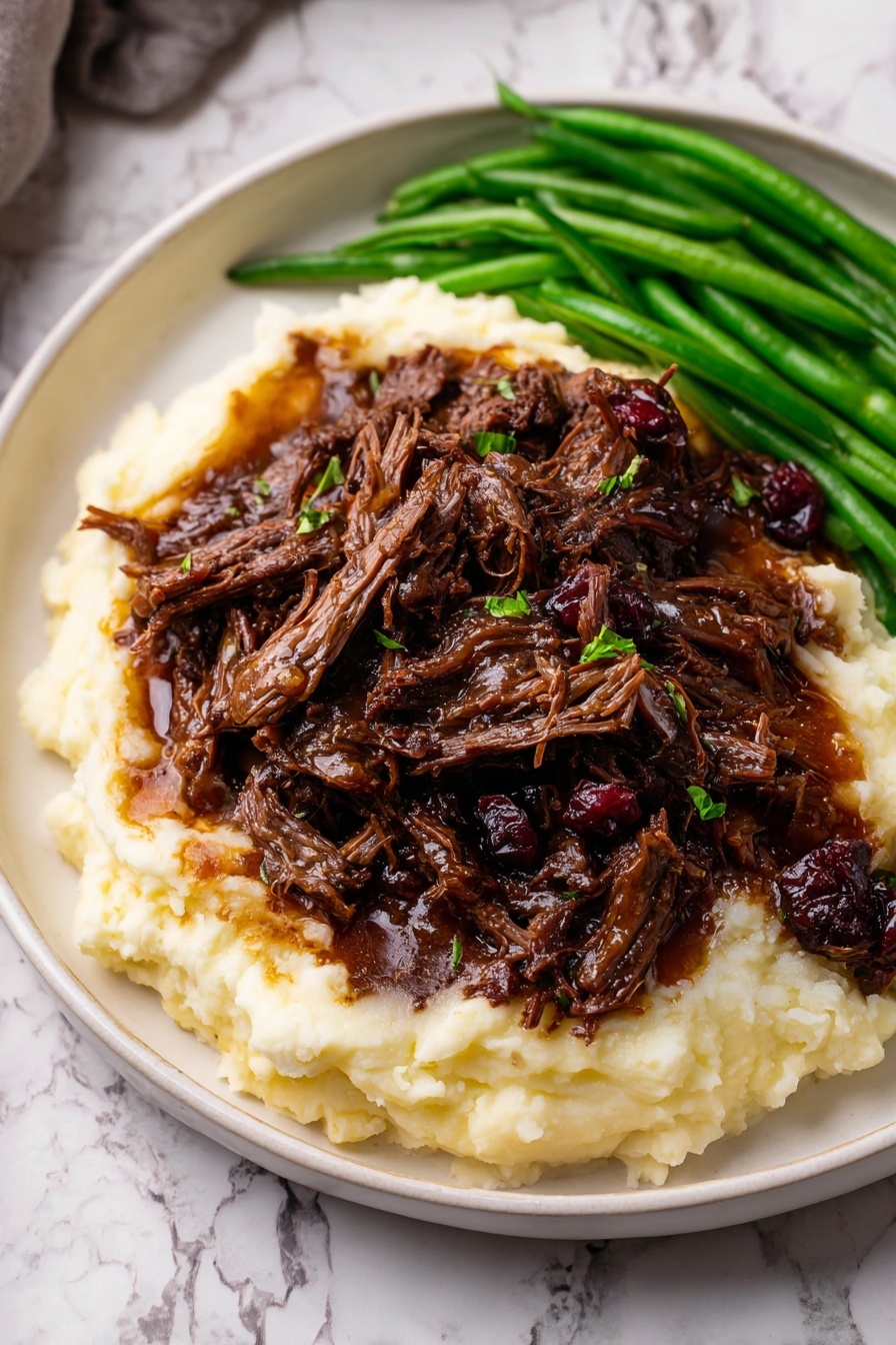 A white plate with ridged edges holds three layers. The bottom layer is creamy, smooth mashed potatoes in a soft off-white color, spread thick and slightly rounded. On top, a generous pile of dark brown shredded beef, moist and tender with visible strands and small chunks, sits in the center, with a few pieces showing a rich, reddish-brown hue. To the side, bright green cooked green beans add a fresh pop of color. The plate is set on a white marbled surface. Photo taken with an iphone --ar 2:3 --v 7