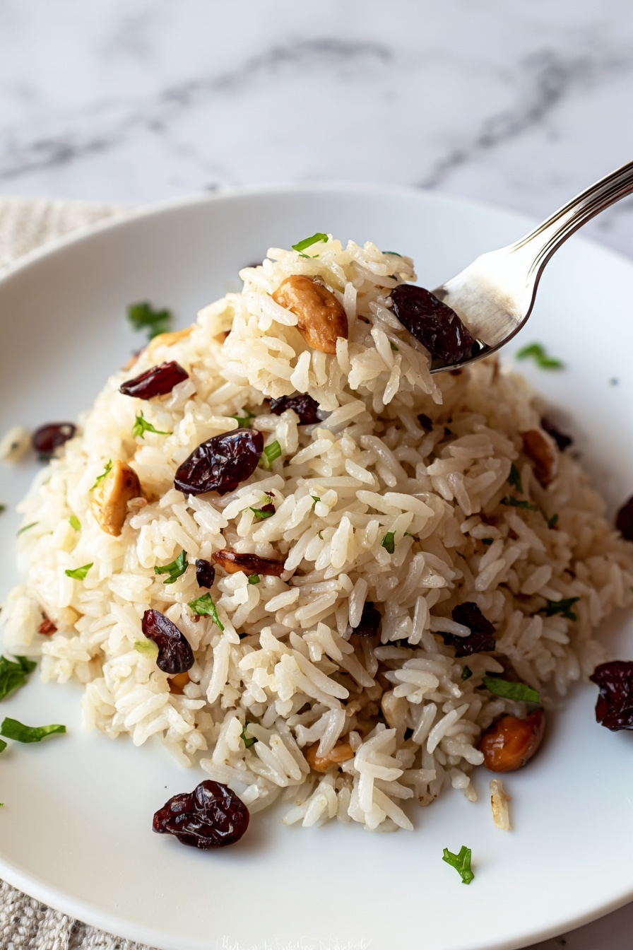 A white plate holds a mound of rice pilaf with visible layers of light brown cooked rice mixed with dark red cranberries and light brown almond slices. Small green herb pieces are scattered throughout and around the base of the rice, adding a fresh touch. The background shows a cozy, soft texture with some out-of-focus red and green elements. A silver fork is partially visible on the left side of the plate. The photo is taken with a white marbled texture underneath. Photo taken with an iphone --ar 2:3 --v 7