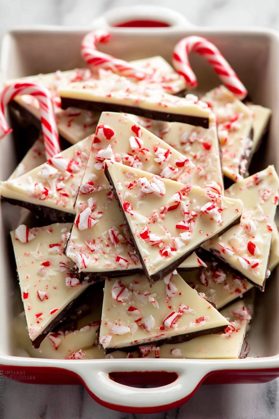 The image shows a white dish with red patterns filled with square and triangular pieces of peppermint bark. Each piece has three layers: a bottom dark chocolate layer, a thin middle layer, and a thick white chocolate top layer sprinkled with small red and white crushed peppermint candy bits. Two whole mini candy canes rest on top of the peppermint bark pieces. The dish sits on a white marbled surface. photo taken with an iphone --ar 2:3 --v 7