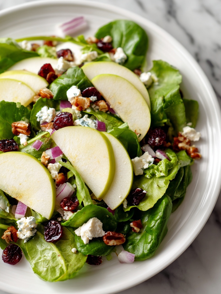 A white plate on a white marbled surface holds a fresh green salad with two metal forks resting on the left side of the plate. The salad has several layers: the base is a mix of dark and light green leafy spinach and lettuce; on top, there are thin slices of bright green apple arranged unevenly; scattered across are small chunks of white cheese and bits of red onion adding purple color; dried cranberries and walnut pieces provide deep red and brown spots throughout the salad. The textures range from smooth apple slices to crumbled cheese and crunchy nuts. Photo taken with an iphone --ar 2:3 --v 7