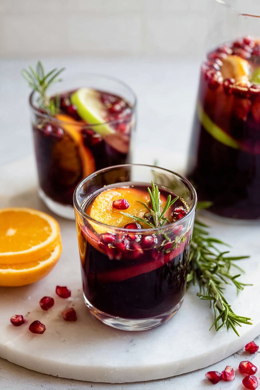 Two clear glasses filled with dark red liquid are placed on a white marbled surface. The front glass shows layers starting with the red drink, floating dark red pomegranate seeds, a thin slice of bright orange, a slice of light green apple, and a sprig of fresh green rosemary, all inside the glass near the rim. The second glass in the background also has similar fruit slices and pomegranate seeds in the dark red liquid. Scattered pomegranate seeds and a small rosemary branch lie beside the glasses on the white marbled surface. photo taken with an iphone --ar 2:3 --v 7