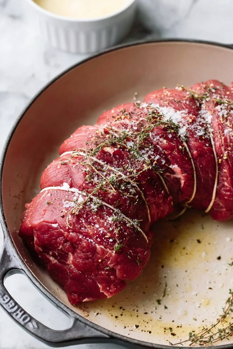 The image shows a thick piece of raw meat tied with string in an oval shape, placed in a white cast iron pan with a black interior. The meat is covered with a mix of light green and white dried herbs and spices scattered evenly on top. The pan is set on a white marbled surface with part of a white bowl holding a light-colored sauce visible near the bottom right corner. The overall scene is brightly lit, focusing on the texture of the meat and the details of the seasoning. photo taken with an iphone --ar 2:3 --v 7