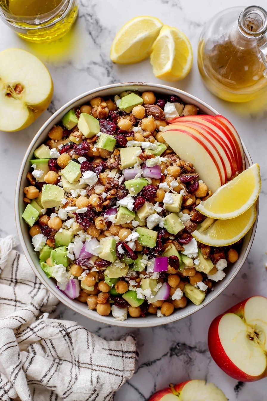 A white bowl filled with a colorful chickpea salad is set on a white marbled surface. The salad has several layers: the base is light brown chickpeas, mixed with bright green chunks of avocado and red apple pieces with their skin on, scattered throughout. There are small purple-red onion cubes and dark reddish dried cranberries spread evenly. White crumbly cheese dots the top, and small bits of brown pecans add texture. On the side of the bowl, there are three lemon wedges placed neatly. Around the bowl, part of a sliced apple, lemon halves, a glass bottle of olive oil, and a striped white cloth are visible. photo taken with an iphone --ar 2:3 --v 7