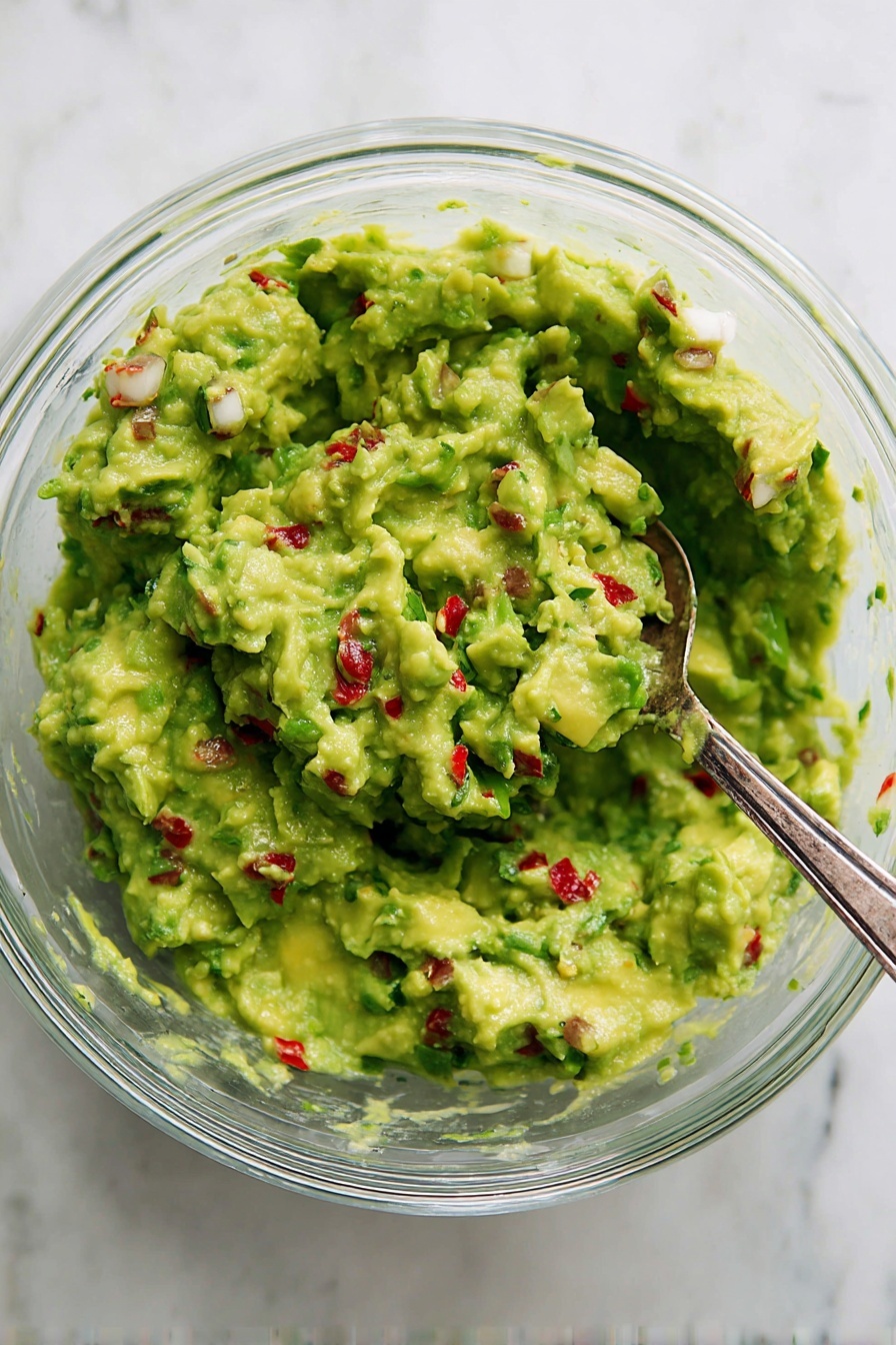 A clear glass bowl filled with bright green guacamole that has a chunky texture with visible small pieces of avocado and bits of white onion, cilantro, and red chili scattered evenly throughout. There is a silver spoon partially submerged on the right side of the bowl. The bowl sits on a white marbled surface. photo taken with an iphone --ar 2:3 --v 7