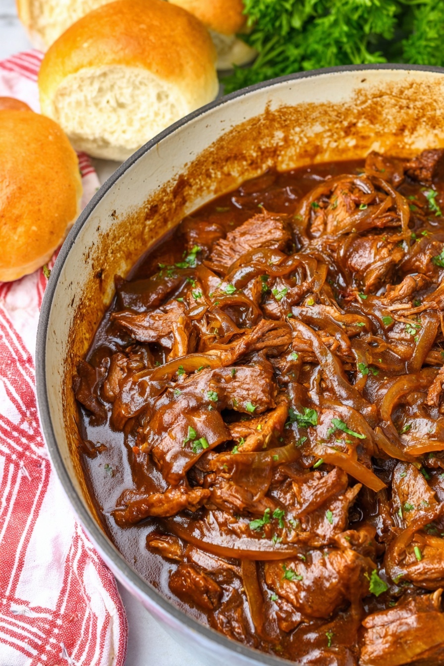 A close-up view of a blue-handled white cast iron pot filled with tender slices and shredded pieces of brown beef mixed with thin light brown onion strips, all coated in a thick dark brown sauce that clings to the meat and edges of the pot. To the upper left of the pot, three soft, round bread rolls with a golden-brown top crust sit on a red and white striped cloth over a white marbled surface, creating a cozy, hearty meal setting. photo taken with an iphone --ar 2:3 --v 7