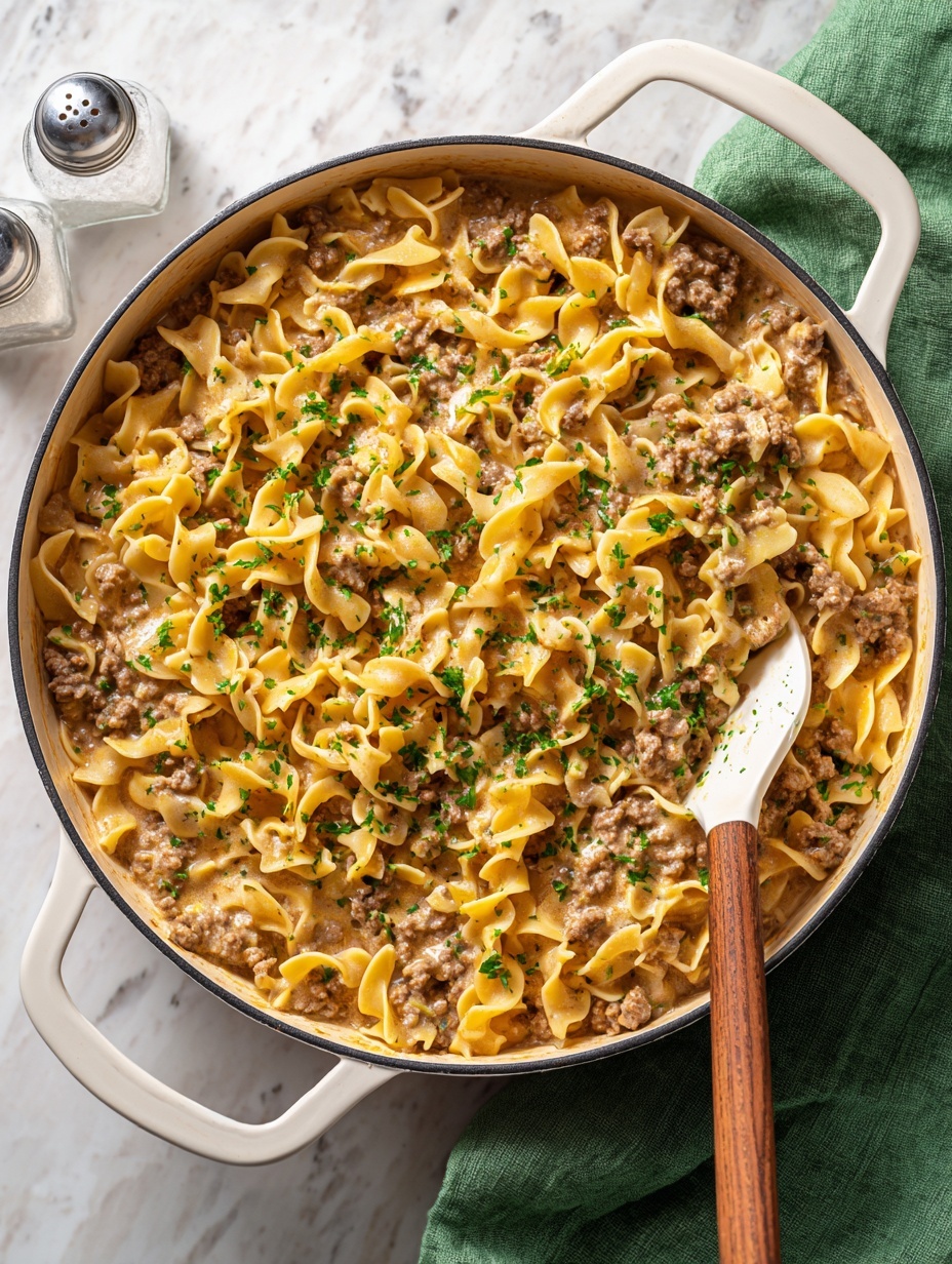 A large white pan filled with creamy beef and egg noodles, showing a mix of light beige egg noodles and brown cooked ground beef pieces all coated in a thick, creamy sauce. The dish is topped with small pieces of chopped green parsley scattered evenly over the noodles and beef. A white spatula with a wooden handle is partially inside the pan, scooping some of the noodles and beef. The pan sits on a white marbled surface, with a green and white patterned cloth partially visible at the bottom. Nearby on the surface is a small clear salt shaker. photo taken with an iphone --ar 2:3 --v 7