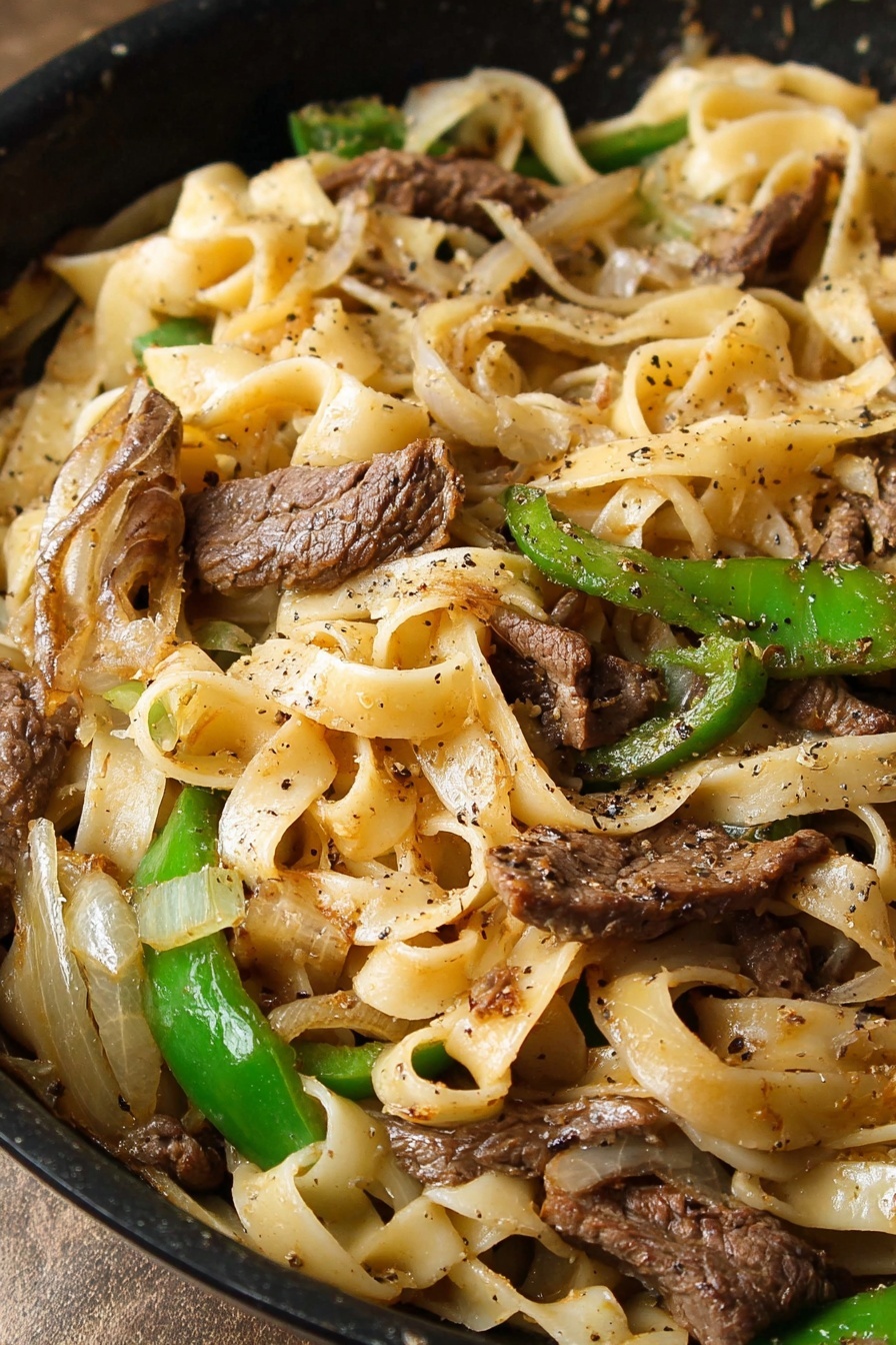 The image shows a close-up of a black pan filled with wide, flat noodles that are creamy light yellow in color and soft in texture. On top of the noodles, there are thin strips of cooked brown beef that have a slightly crispy edge. Scattered among the beef and noodles are slices of green bell pepper and translucent cooked onions with light brown caramelized spots. The dish is lightly seasoned with black pepper sprinkled across the layers. The pan sits on a white marbled surface with a striped cloth partially visible under the pan. photo taken with an iphone --ar 2:3 --v 7