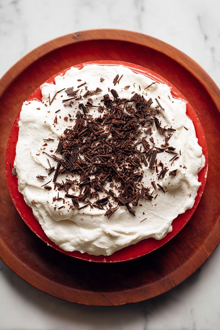 The image shows a three-layered dessert on a wooden stand with a white marbled surface underneath. The bottom layer is a dark, almost black crumbly base, likely chocolate. The middle layer is thick and smooth with a bright red color, creating a strong contrast with the bottom. On top of the red layer, there is a thick, fluffy layer of white cream with a soft texture. Scattered on the cream are small shavings of dark chocolate, adding a touch of texture and decoration. Photo taken with an iphone --ar 2:3 --v 7
