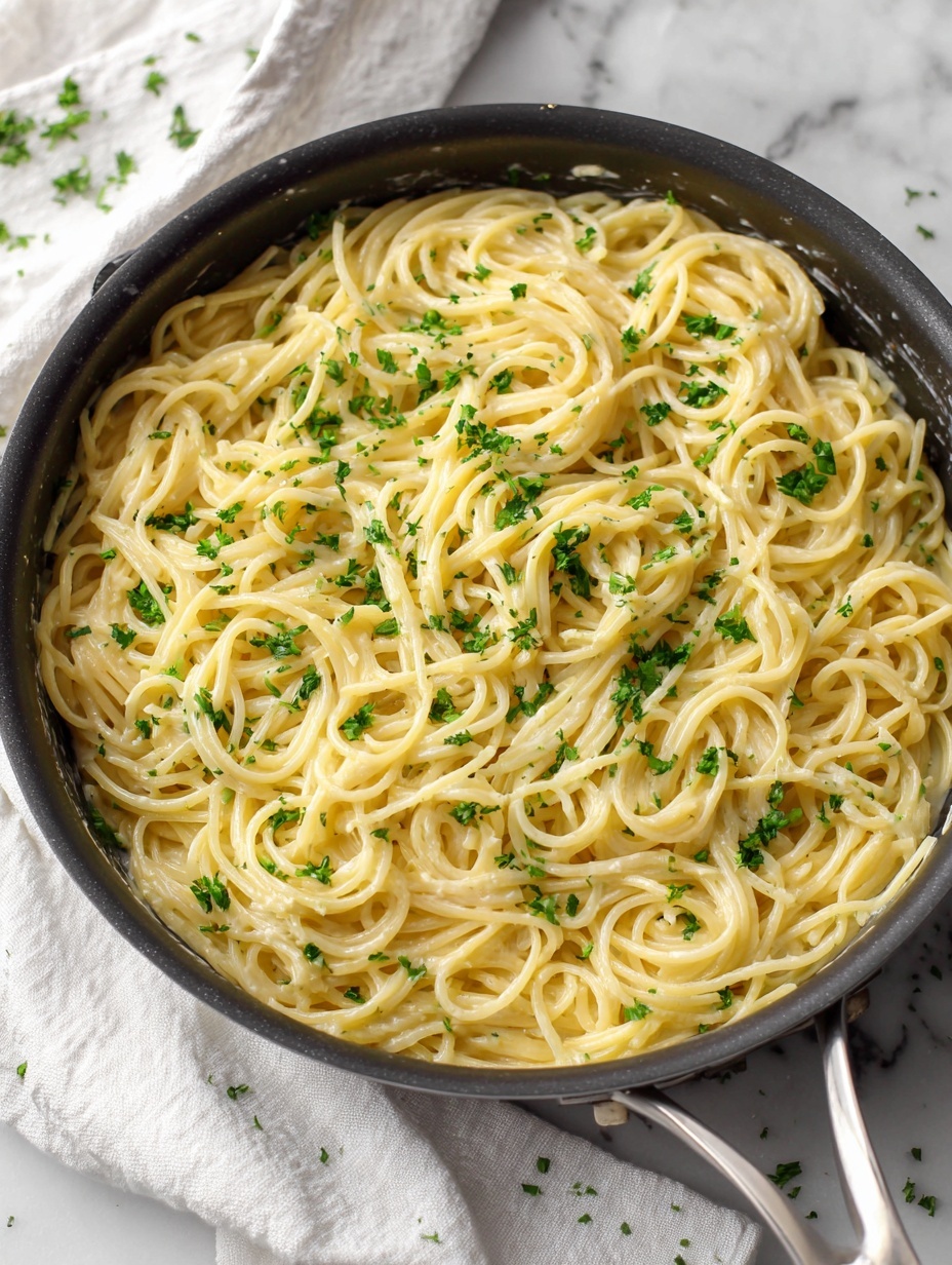 A large round silver pan filled with a big pile of light yellow spaghetti noodles, mixed with small green herb pieces scattered evenly throughout. Two silver forks are twirling some of the noodles together near the center of the pan, showing the smooth and creamy texture of the pasta. The pan sits on a white marbled surface with a soft white cloth partly visible under it. The lighting is soft and natural, highlighting the shiny noodles and the fresh green herbs. photo taken with an iphone --ar 2:3 --v 7