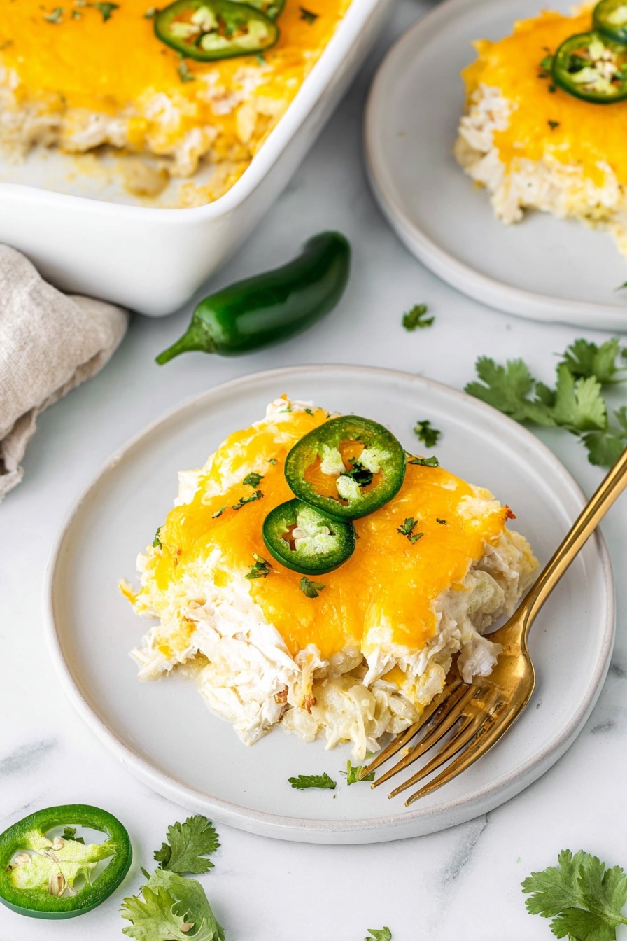 A square piece of cheesy casserole sits on a white plate, topped with melted bright yellow cheddar cheese and two green jalapeño slices, with small bits of green herbs sprinkled over it. The dish has multiple layers including a creamy white sauce and shredded chicken visible underneath the cheese. A gold fork rests beside the casserole on the plate. The casserole is part of a larger white baking dish shown in the top right corner with a fresh green cilantro sprig nearby on a white marbled surface. Photo taken with an iphone --ar 2:3 --v 7